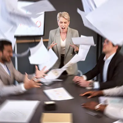 Upset business people at a conference table with papers flying through the air around them.