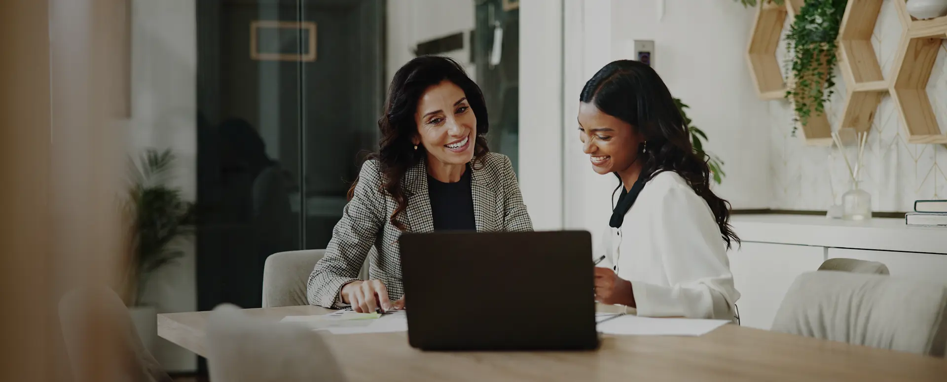 Two women sitting at a table and smiling as they review information on papers and a laptop.