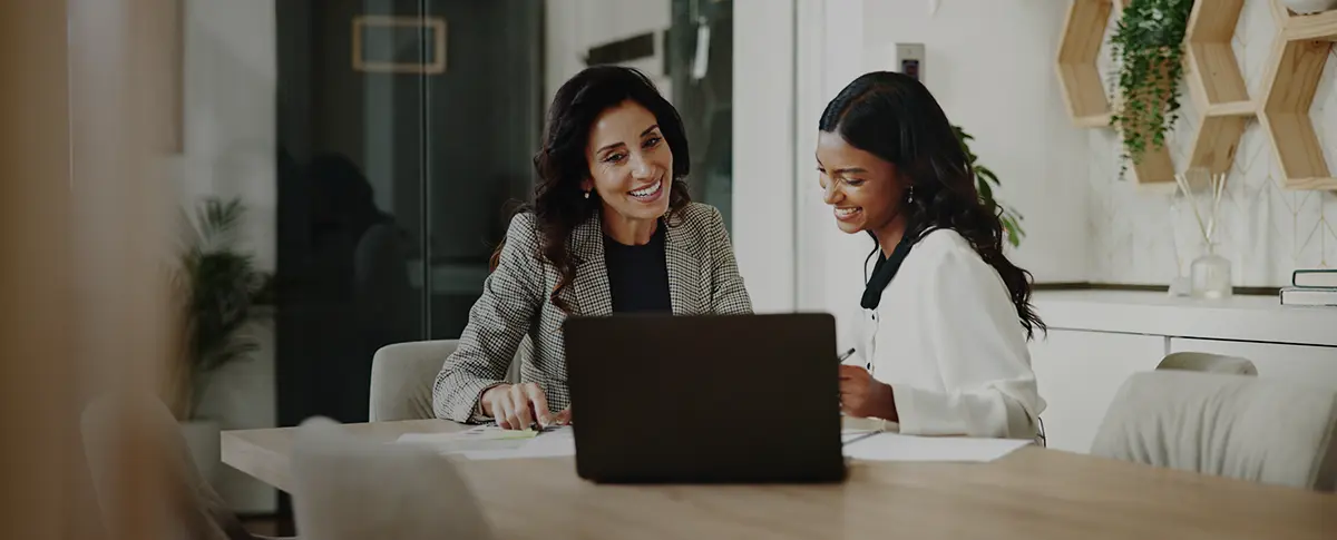 Two women sitting at a table and smiling as they review information on papers and a laptop.