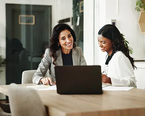 Two women sitting at a table and smiling as they review information on papers and a laptop.