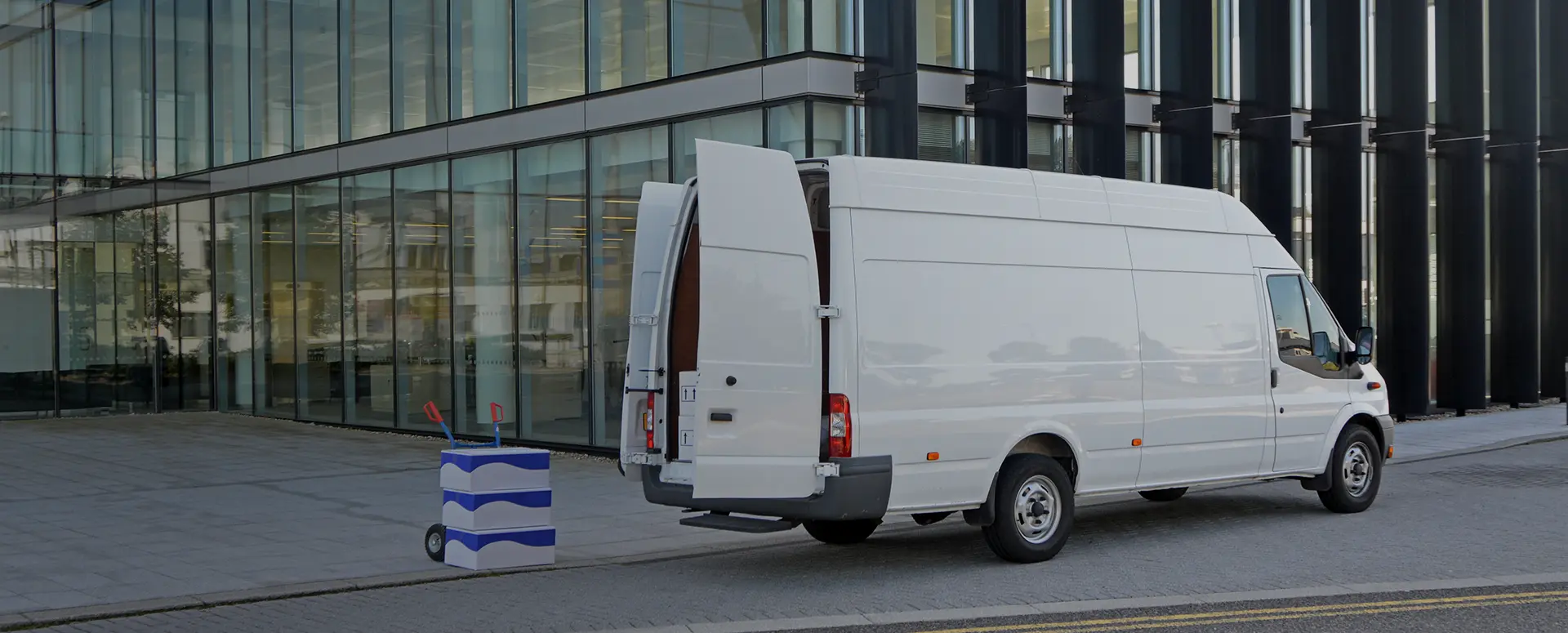 A white delivery van parked with its back doors open and a stack of boxes on a dolly sitting behind it.