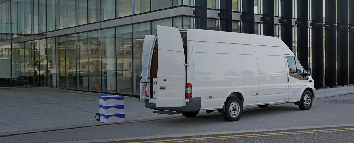 A white delivery van parked with its back doors open and a stack of boxes on a dolly sitting behind it.