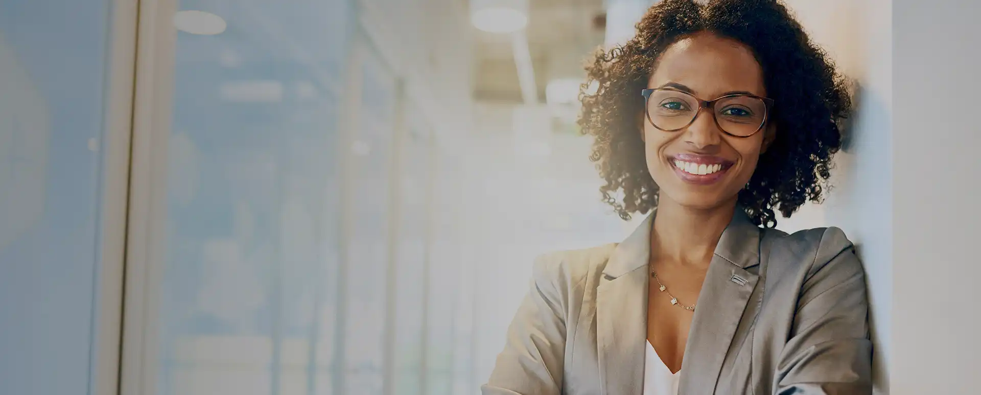 A woman wearing a grey jacket and glasses smiling as she leans against a wall.