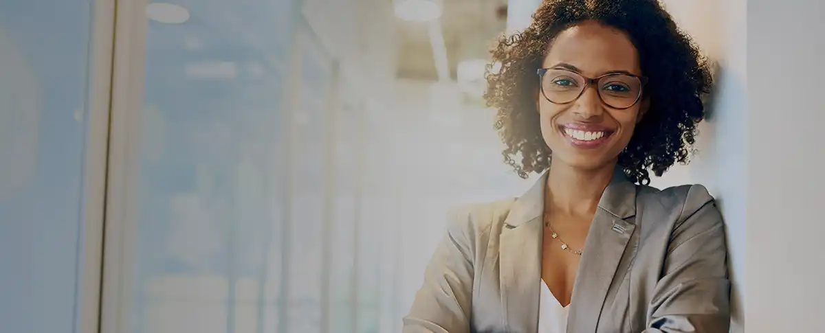 A woman wearing a grey jacket and glasses smiling as she leans against a wall.