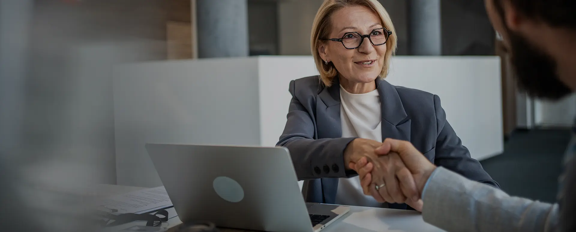 A woman with glasses sitting at a table with a laptop and shaking the hand of a man across form her.