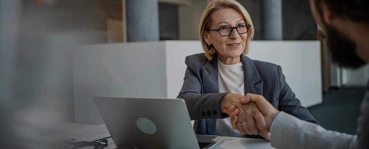 A woman with glasses sitting at a table with a laptop and shaking the hand of a man across form her.