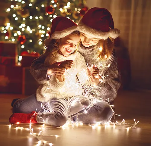 A woman and young girl wearing Santa hats and holding a string of glowing Christmas string lights.
