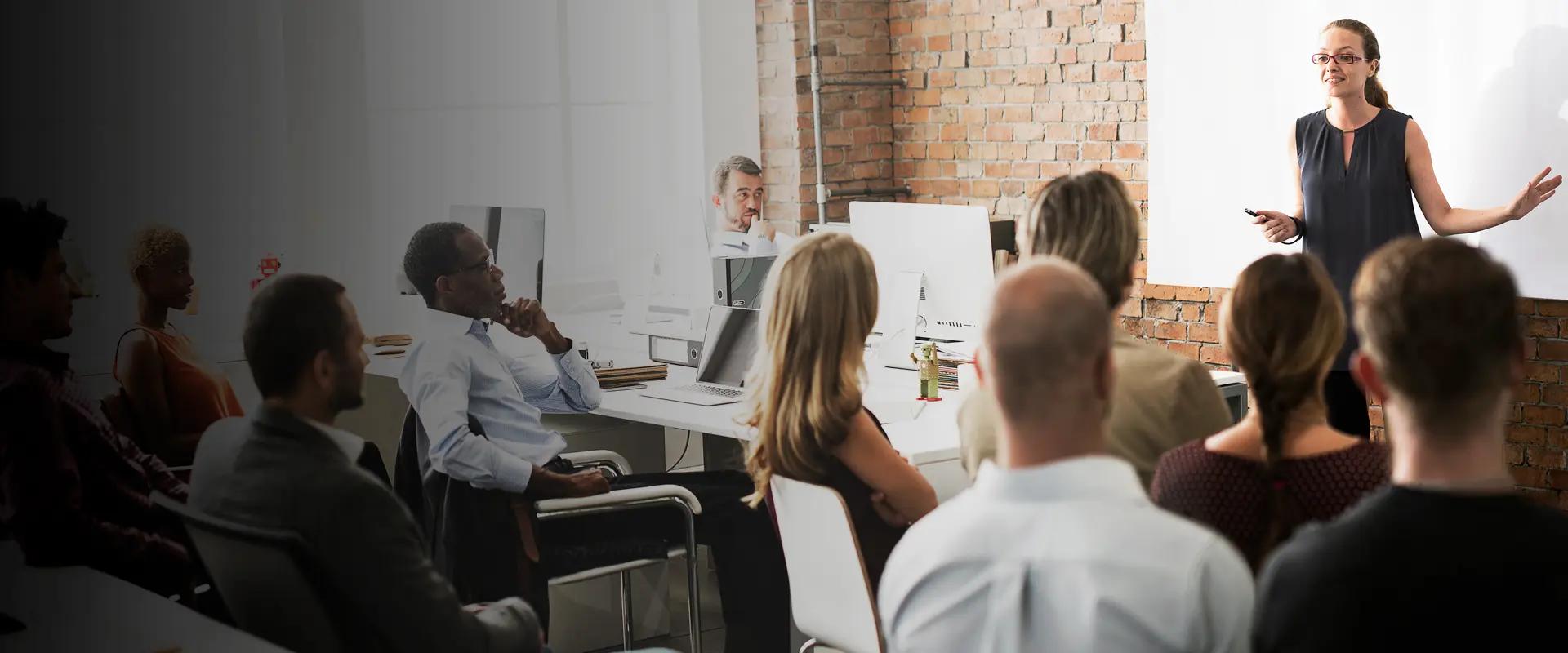A group of people siting in a room with woman standing in front and speaking.
