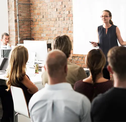 A group of people siting in a room with woman standing in front and speaking.