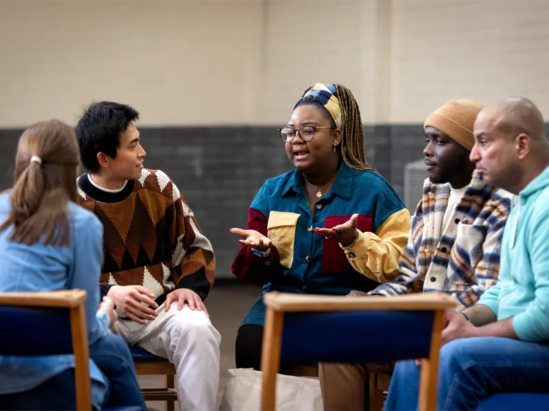 A group of people sitting in a circle of chairs listening to one member talk.