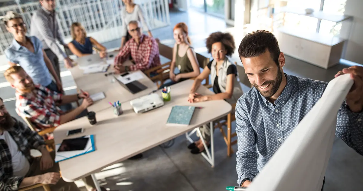 A meeting of people in people sitting at a couple tables with a man writing on a dry erase baord.