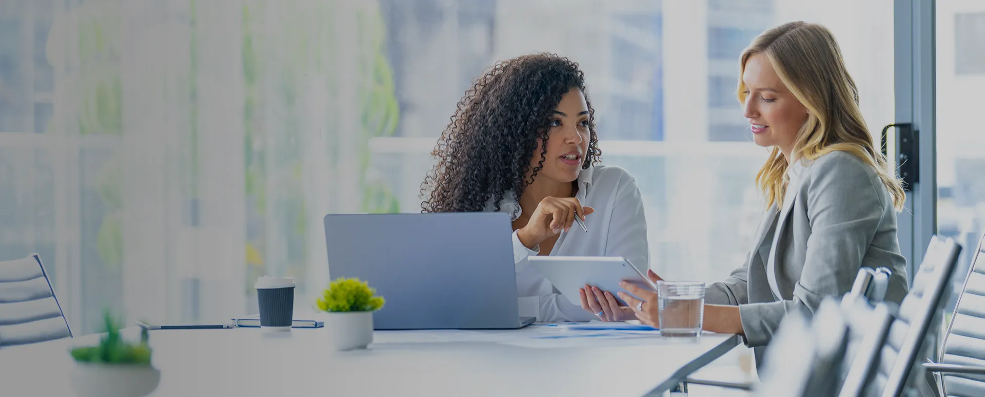 Two women site a conference table reviewing information on a laptop and tablet.