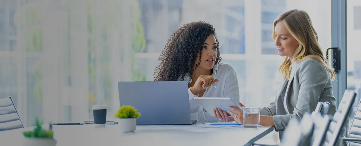 Two women site a conference table reviewing information on a laptop and tablet.