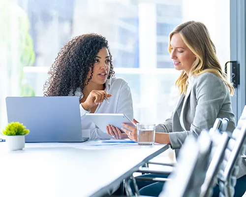Two women site a conference table reviewing information on a laptop and tablet.