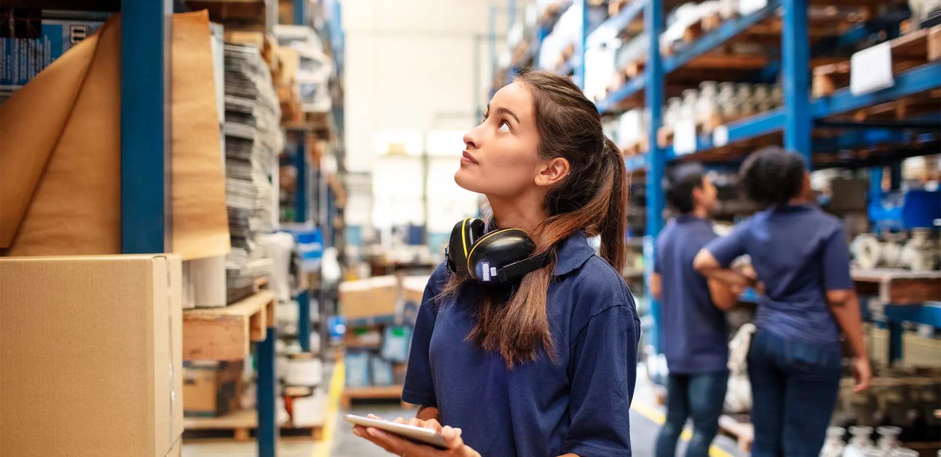 A worker in a warehouse surveying inventory while carrying a tablet.