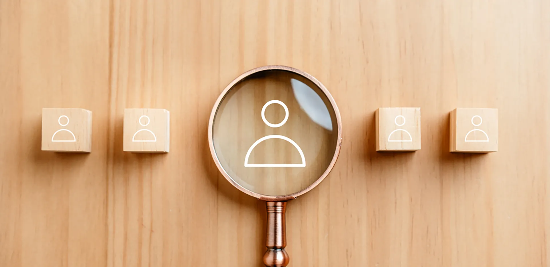 A magnifying glass on an icon of a person against a wood background.