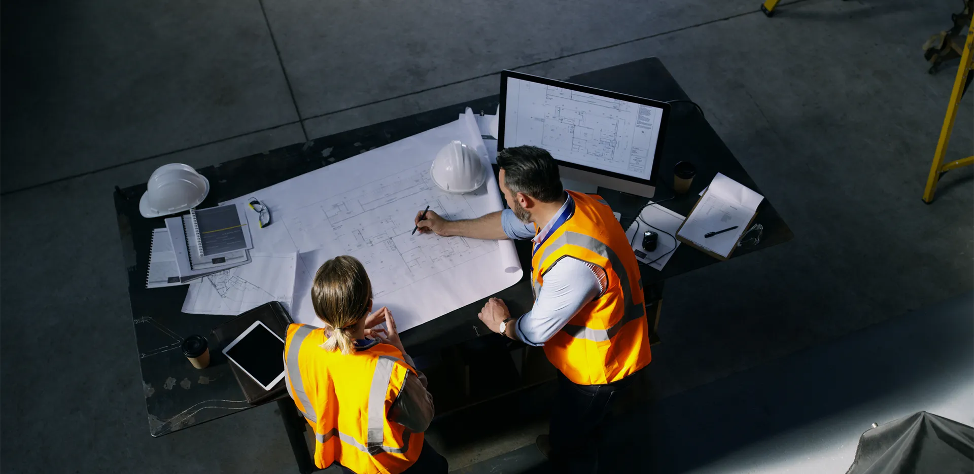 Two contractors in orange safety vests reviewing blueprints on a large desk.