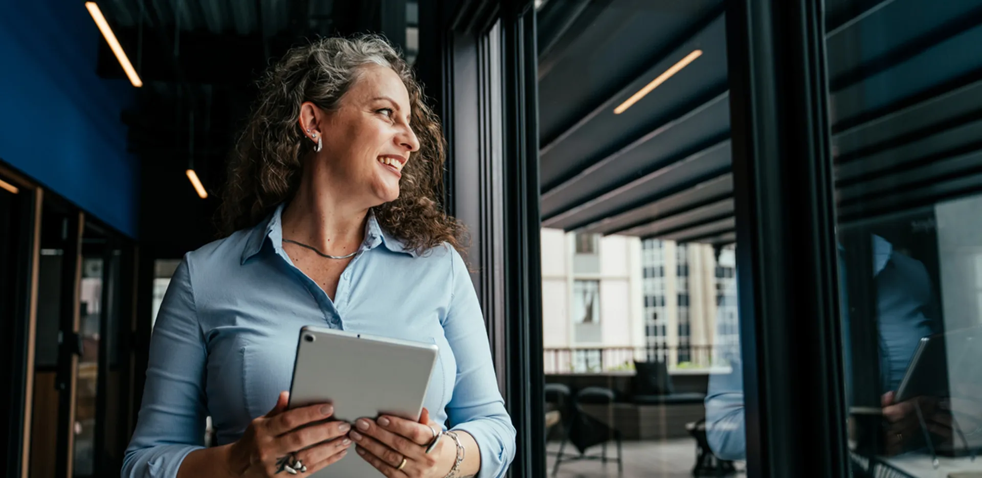 A smiling woman holding a tablet and looking across an office space.