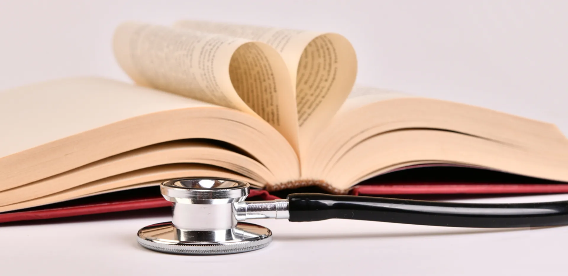 A stethoscope sitting on a white surface in front of an open book with two pages folded like a heart.