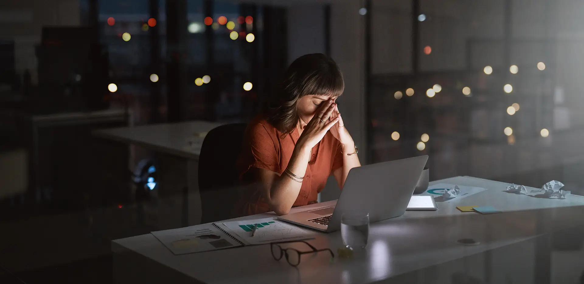 A woman sitting at her desk late at night, looking at a laptop computer.