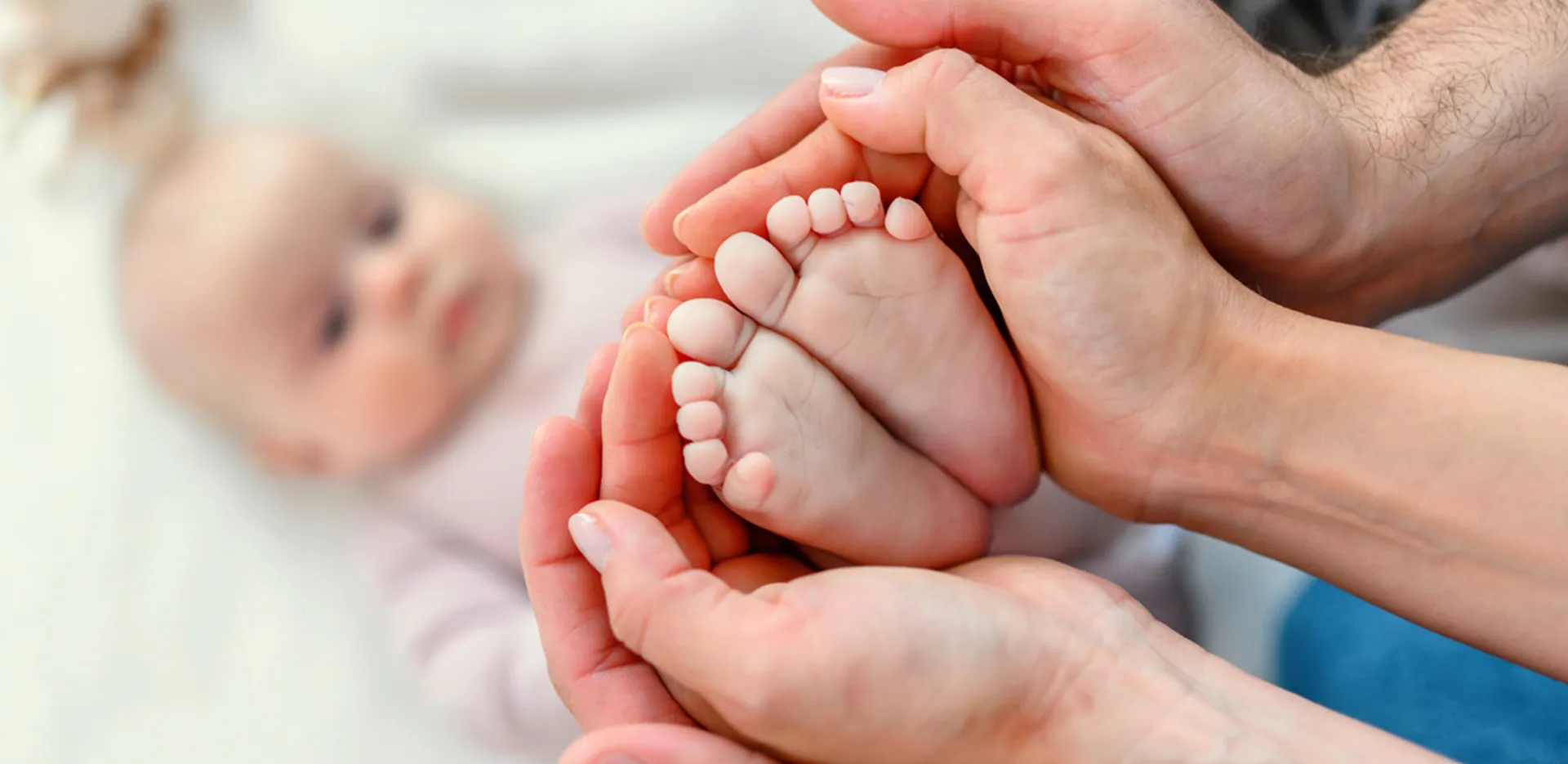 A baby's feet being cupped by two pairs of hands. An out-of-focus baby is in the background.