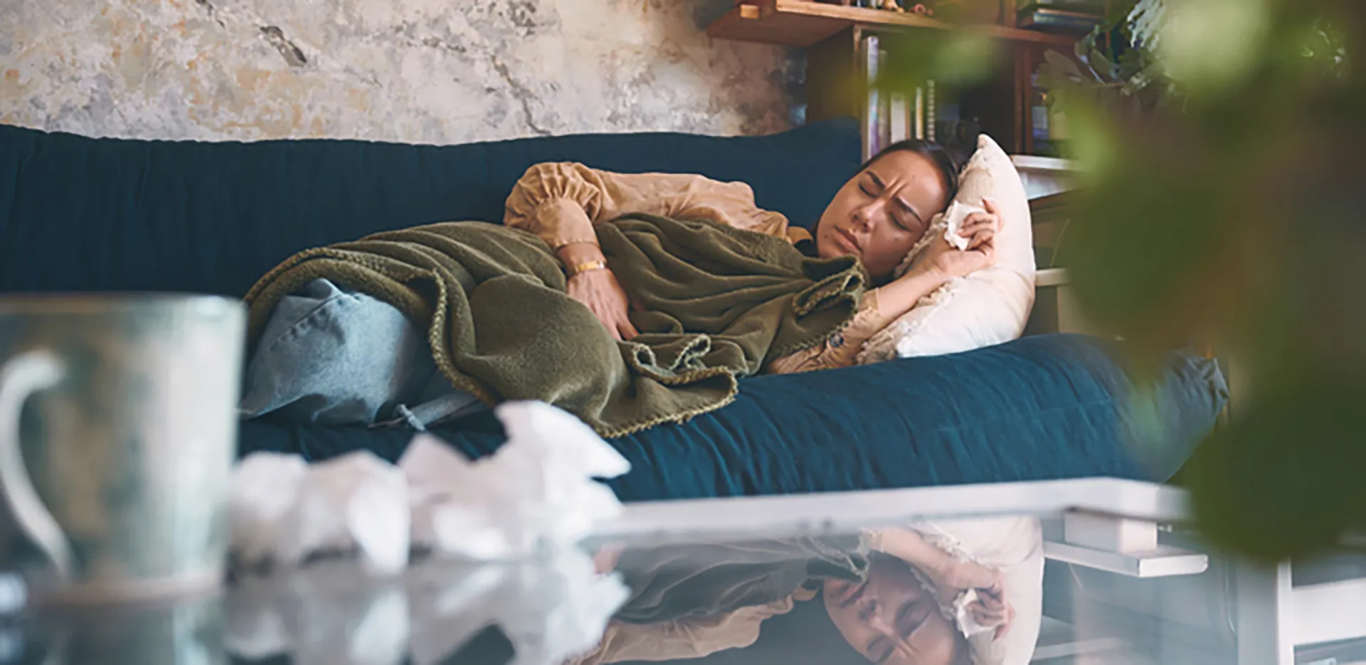 A person on their couch taking a nap. Tissues and a mug of tea can be seen in the foreground.