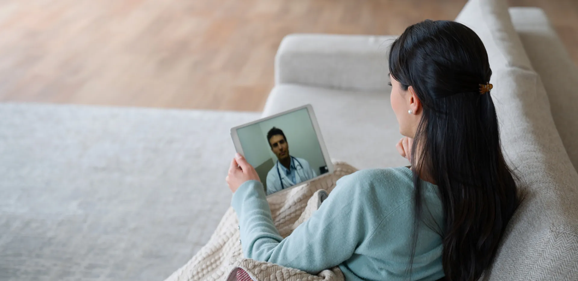 A woman in a blue sweater sitting on w white couch hold a tablet in her hand while on a telehealth call with a doctor.