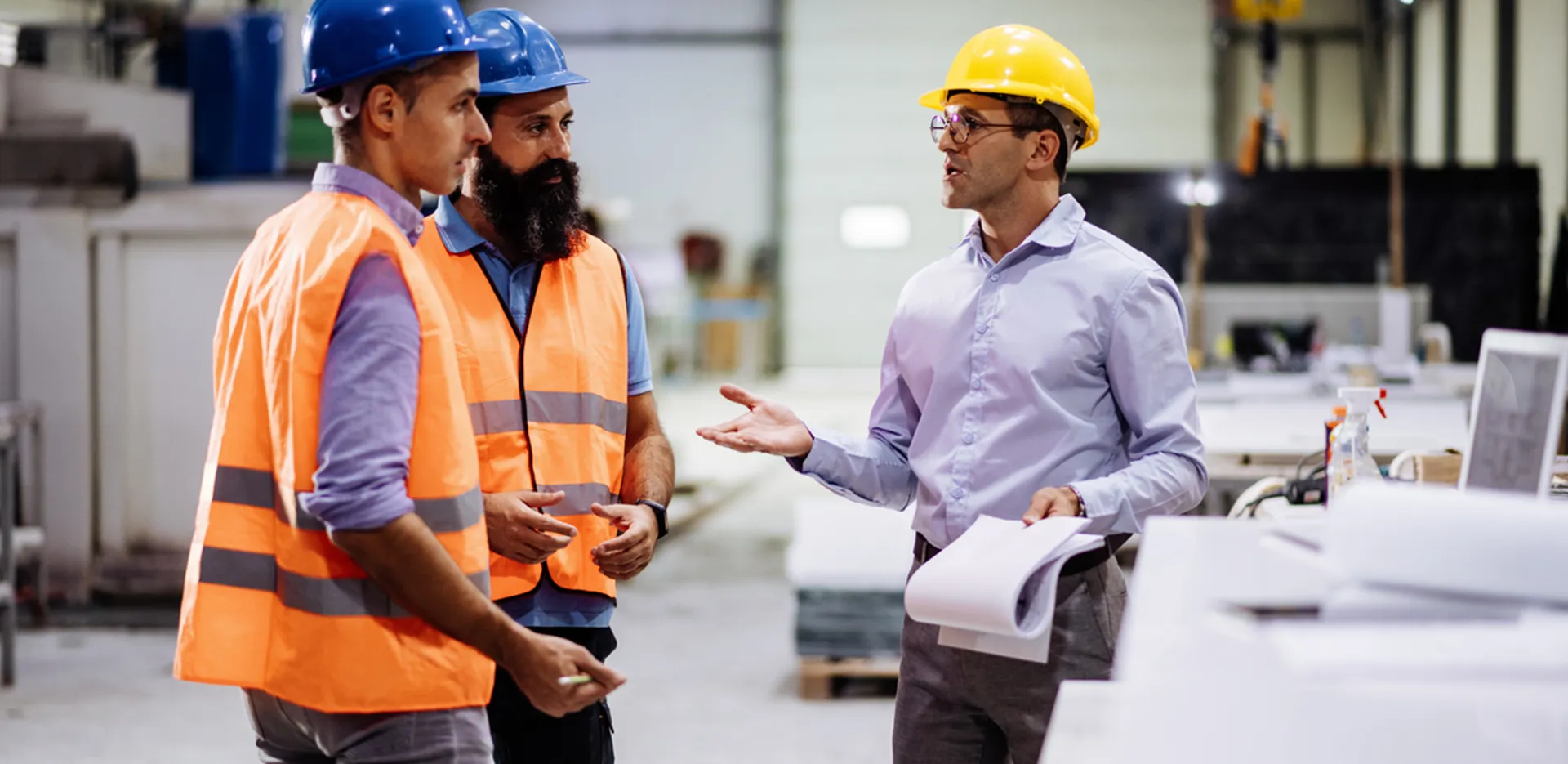 Three men in hard hats and safety gear having a discussion in an industrial space.