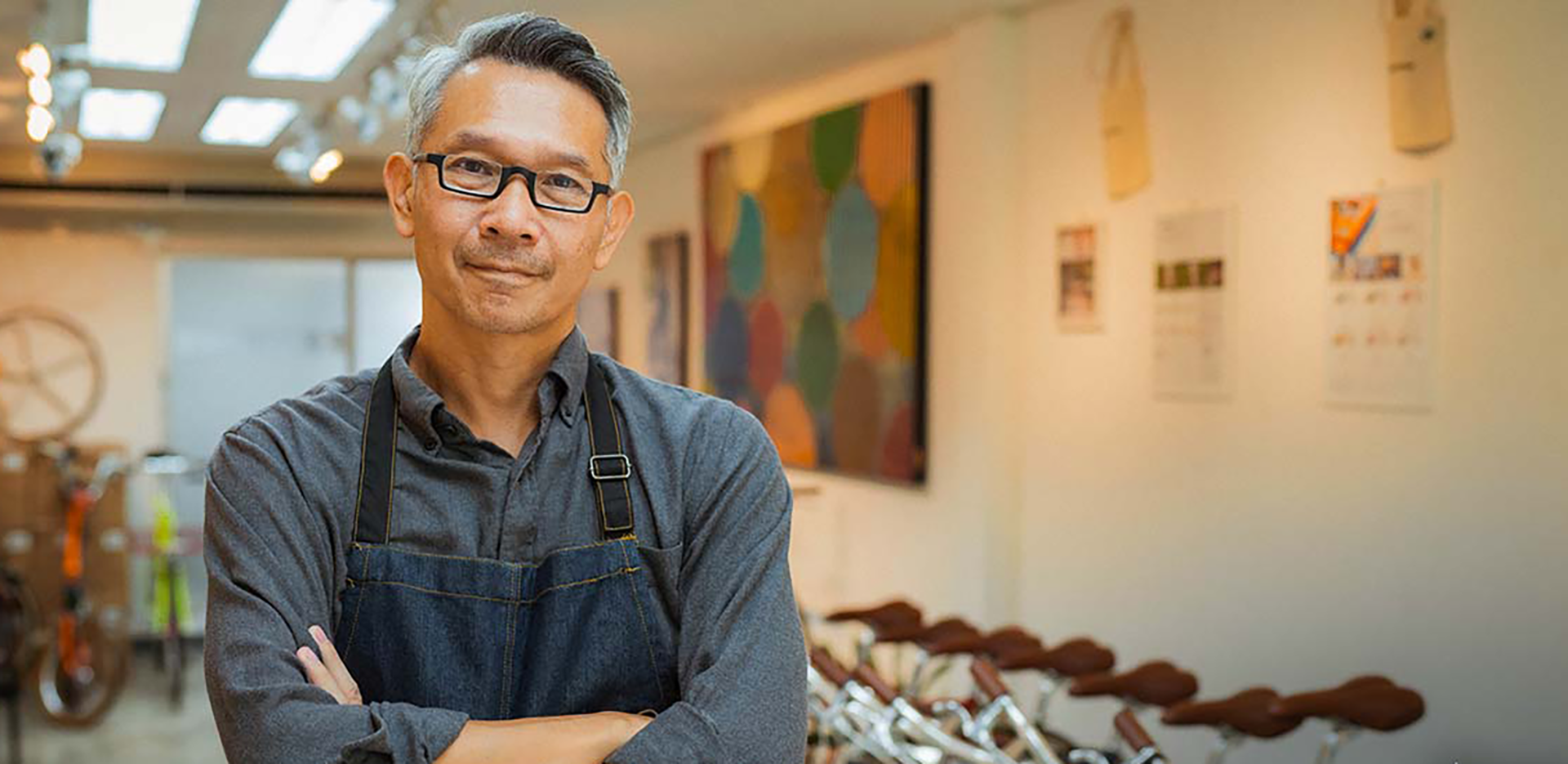 A bike shop owner wearing an apron looking at the camera with his arms crossed.