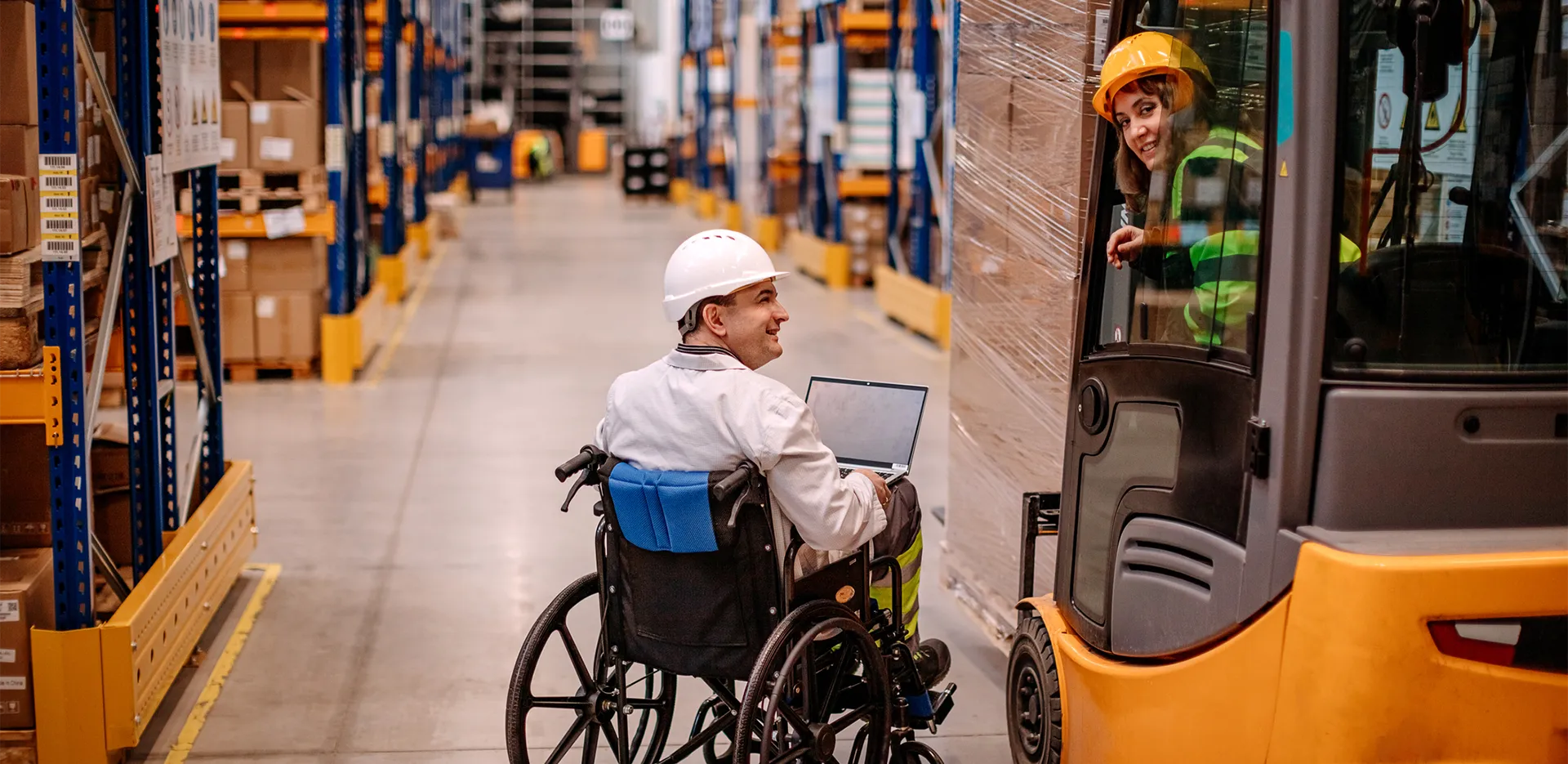 A worker using a wheelchair speaks with another worker driving a forklift. Both are wearing hard hats.