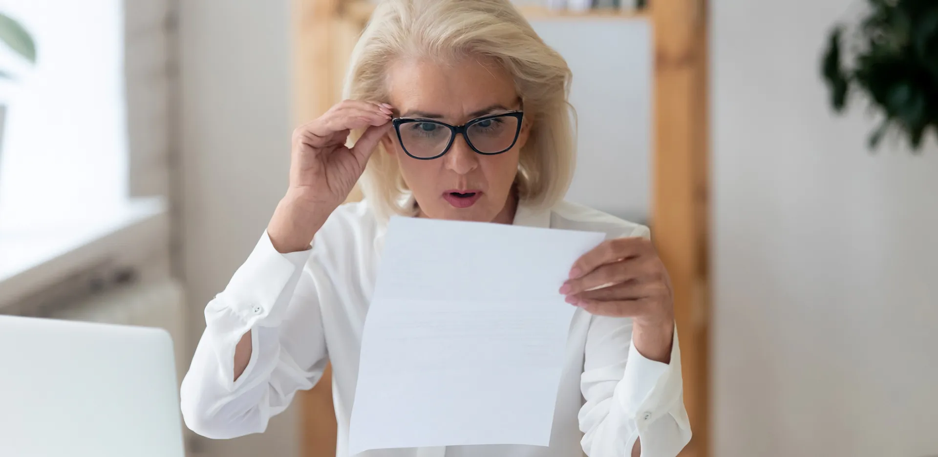 A woman holds her glasses and she reads a piece of paper in disbelief.