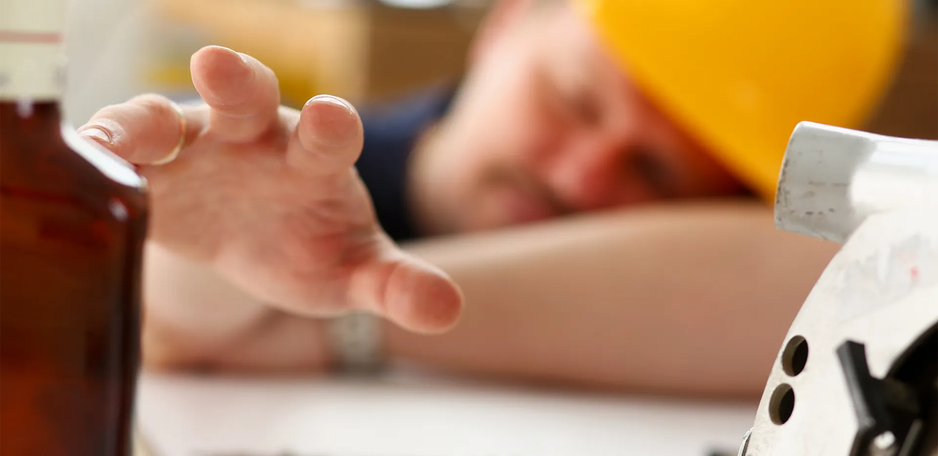 An out-of-focus construction worker wearing a hard hat reaches towards a brown bottle.