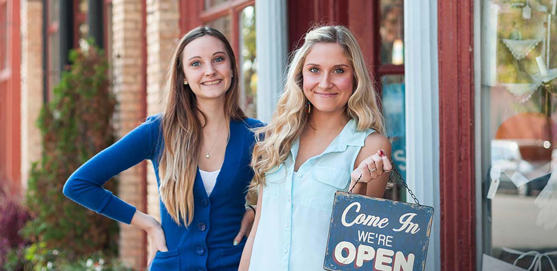 Two small business owners pose in front of their business holding a 'Come in we're open' sign.