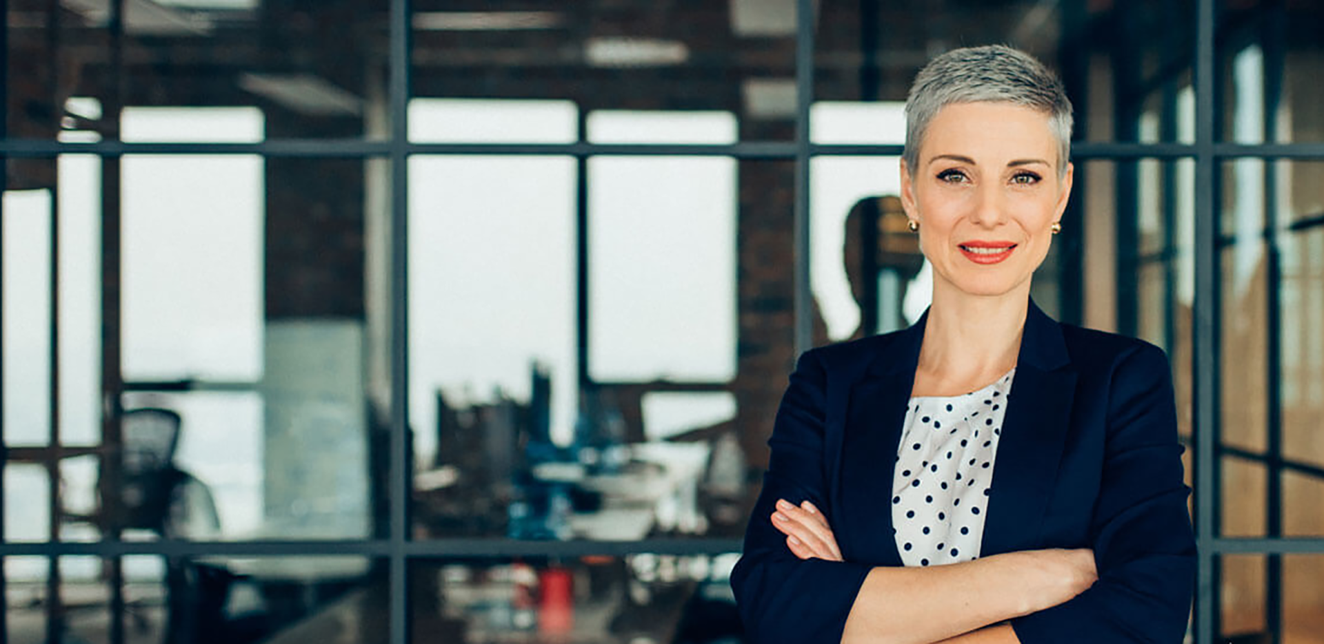 A professionally dressed woman standing with her arms folded in front of a glass window.