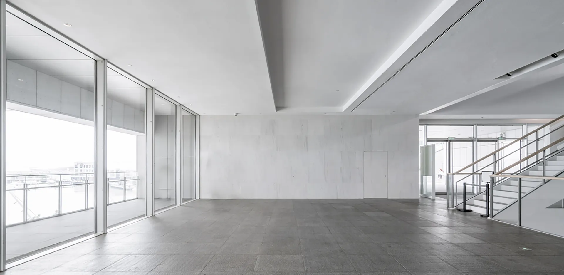 The concrete interior of a vacant building with large windows and stairs.
