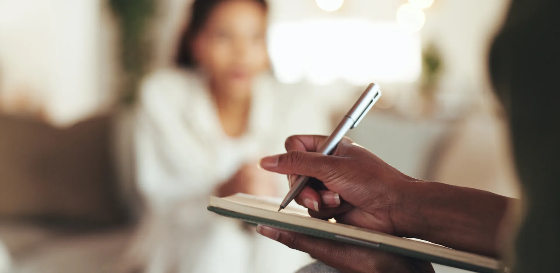 A hand writing in a notebook with the blurry shape of a woman in the background.