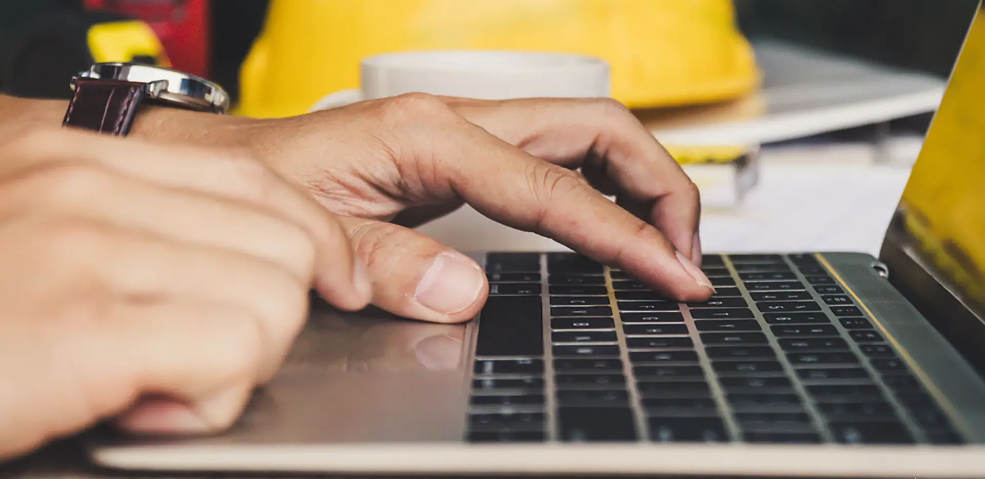 A person working on a laptop computer with a hard hat in the background.