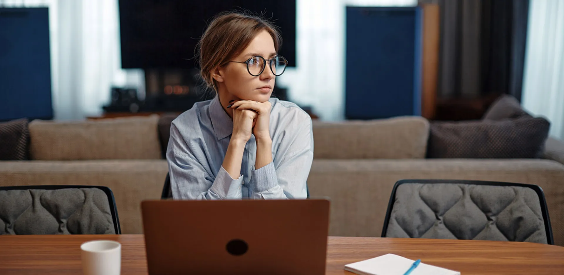 A young woman sitting at a table with a laptop, staring away in the distance.