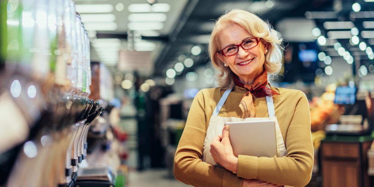 Portrait of happy senior woman holding digital tablet, standing between shelves with food and enjoying her job