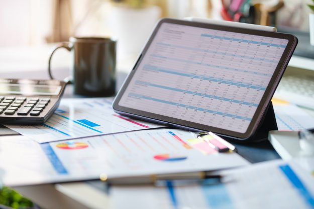 Digital tablet with profit and tables on screen, calculator, financial reports on wooden desk.