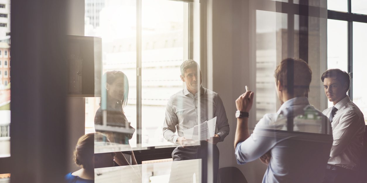 A photo of business people having discussion in board room. Male and female professionals are seen through glass. They are working in office.