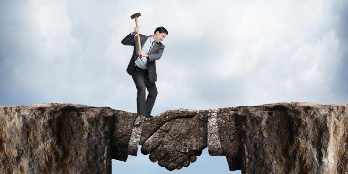 A businessman swings a sledgehammer at a land bridge connecting two cliffs as he tries to chip away at the metaphorical agreement represented by the bridge formed in the shape of a handshake.